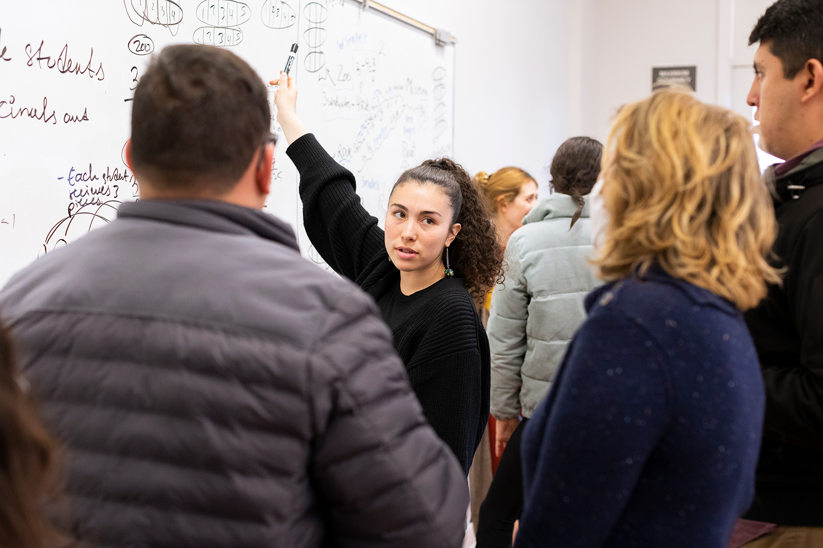 person gesturing to a whiteboard while speaking to several people