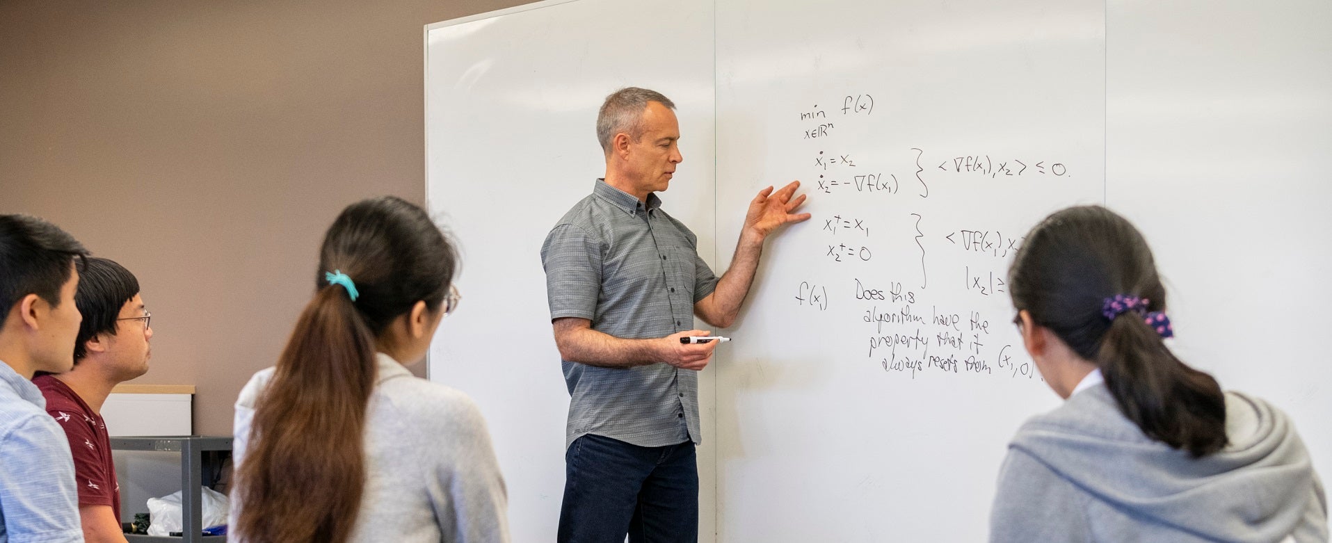 person gesturing at an equation on a whiteboard in front of four seated people